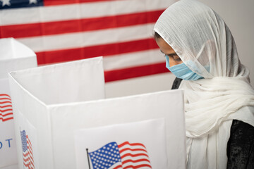 Girl with Hijab or head covering and mask worn busy at polling booth with US flag as background -...