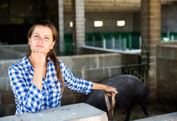 Portrait of thoughtful young female farmer standing in stall with black Iberian pig
