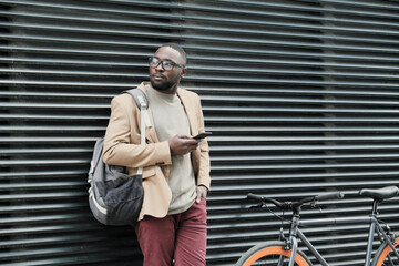African businessman with backpack using his mobile phone while waiting for his colleague in the city outdoors
