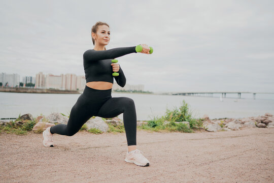 A Woman Of European Appearance, Playing Sports On The Street, Doing Exercises Squat Lunges Forward, Holding Small Dumbbells. Looks Ahead Thinks About Proper Nutrition