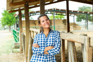 Fototapeta premium Happy confident young woman owner of ranch posing near empty stable on summer day
