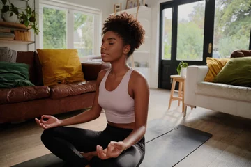 Fototapeten Young black woman doing yoga at home in the lotus position © StratfordProductions