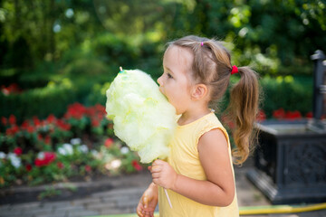 Cute toddler girl eating big yellow candyfloss or cotton candy  in the park on a summer sunny day. 