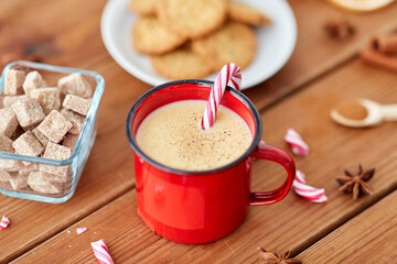 christmas and seasonal drinks concept - red cup of eggnog with candy cane, oatmeal cookies, fir tree branches and sugar on wooden background