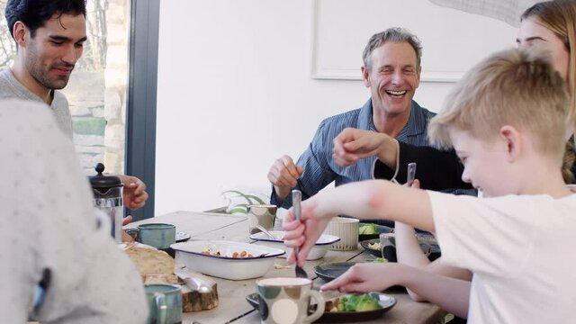 Multi-generation family sitting around table at home in pyjamas enjoying brunch together with grandmother helping granddaughter to cut up food - shot in slow motion
