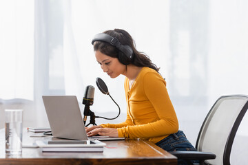 brunette asian announcer in wireless headphones typing on laptop near microphone in studio