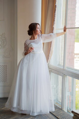 Fototapeta premium Full-length portrait of the bride in a white wedding dress by the window