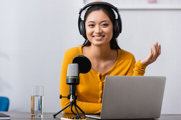 excited asian radio host in wireless headphones looking at camera while sitting near microphone and...