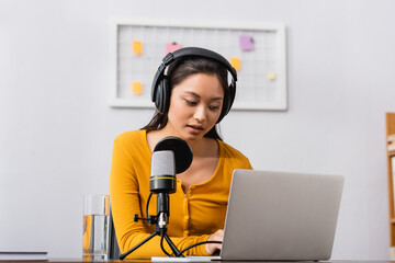 concentrated asian radio host in wireless headphones using laptop near microphone in studio