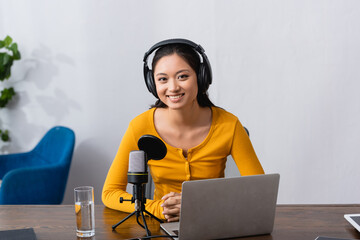 joyful asian announcer in wireless headphones looking at camera while sitting near laptop and...