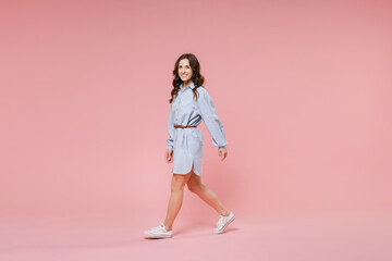 Full length side view portrait of smiling cheerful young brunette woman 20s wearing casual blue shirt dress posing walking going looking camera isolated on pastel pink colour background in studio.