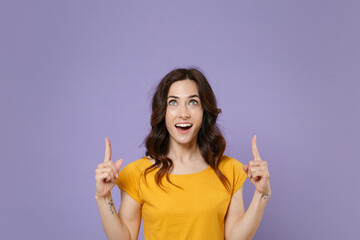 Shocked amazed young brunette woman 20s wearing basic yellow t-shirt posing standing pointing index fingers up on mock up copy space isolated on pastel violet colour background, studio portrait.