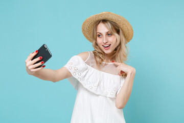 Funny young blonde woman 20s wearing white summer dress hat standing doing selfie shot on mobile phone pointing index finger on herself isolated on blue turquoise colour background studio portrait.