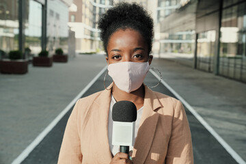 Portrait of African young reporter in protective mask holding microphone and looking at camera while standing in the city - obrazy, fototapety, plakaty