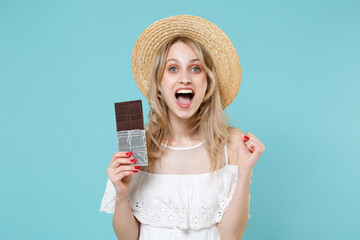 Surprised excited young blonde woman 20s wearing white summer dress hat standing hold in hand chocolate bar clenching fist looking camera isolated on blue turquoise colour background studio portrait.