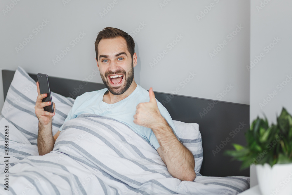 Wall mural excited young bearded man in basic blue t-shirt using mobile cell phone typing sms message showing t