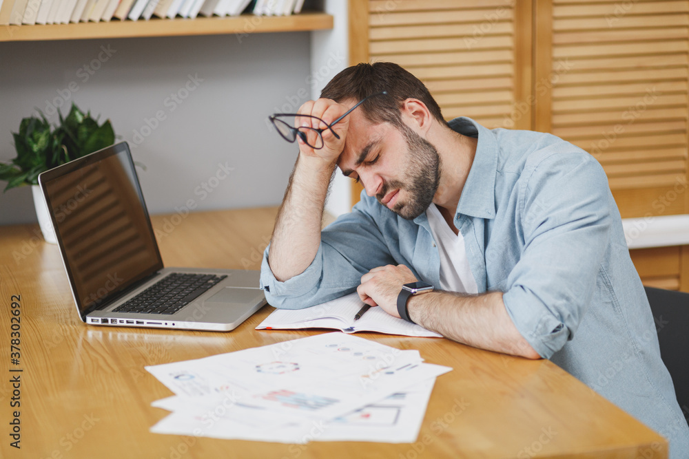 Wall mural displeased tired exhausted young bearded business man in blue shirt glasses sitting at desk with pap