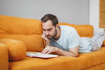 Fototapeta premium Attractive serious handsome young bearded man 20s wearing casual blue t-shirt hold in hands reading book put hand prop up on chin lying on couch resting relaxing spending time in living room at home.