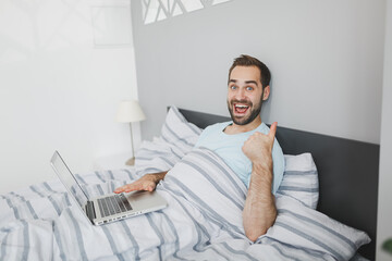 Excited young bearded man 20s in basic blue t-shirt working on laptop pc computer showing thumb up lying in bed with striped sheet pillow blanket resting relaxing spending time in bedroom at home.