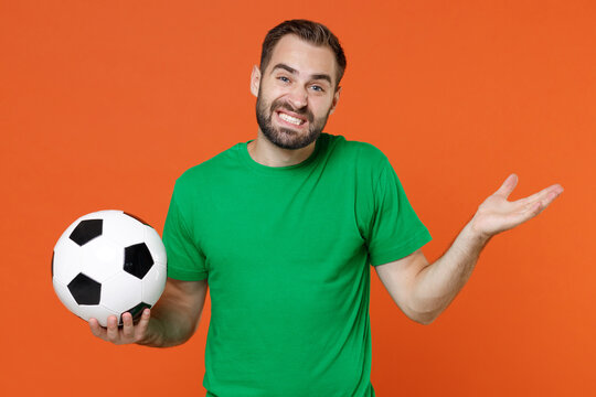 Confused young man football fan in green t-shirt cheer up support favorite team with soccer ball spreading hands isolated on orange background studio portrait. People sport leisure lifestyle concept.