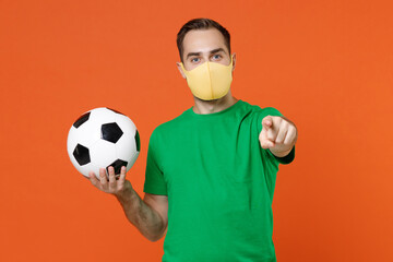 Young man football fan in green t-shirt face mask to safe from coronavirus support favorite team with ball pointing index finger on camera isolated on orange background. People sport leisure concept. - Powered by Adobe