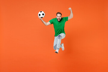 Fototapeta na wymiar Full length portrait of screaming man football fan in green t-shirt cheer up support favorite team with soccer ball jumping clenching fist isolated on orange background. People sport leisure concept.