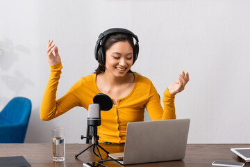excited asian announcer in wireless headphones showing wow gesture near microphone and laptop