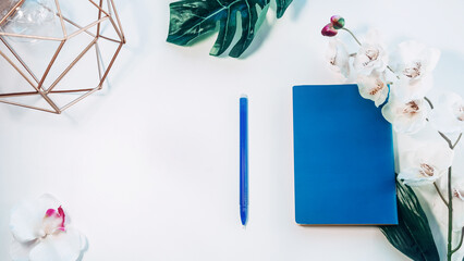 top view of office desk table with digital camera and blue notepad on white background, decorated with orchid and leaves, graphic designer,