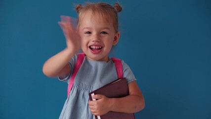 childhood, school, education, upbringing, science concept - close-up happy little blonde caucasian slavic girl with backpack holding book and waving goodbye smiling broadly on solid blue background