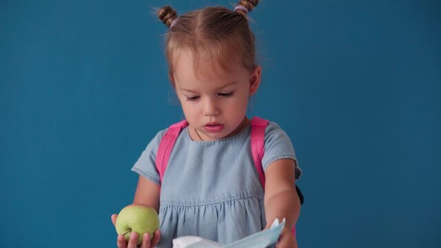 Childhood, School, Education, Upbringing, Science Concept - Close-up Cute Little Blonde Caucasian Slavic Girl With Backpack Unhappy Rip Off And Break Medical Mask Throws Aside On Solid Blue Background