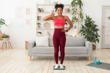 Fototapeta na wymiar African Girl Standing On Weight Scales Gesturing Thumbs Up Indoors