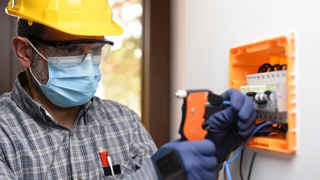 Electrician at work on an electrical panel protected by helmet, safety goggles and gloves; wear the surgical mask to prevent the spread of Coronavirus. Construction industry. Covid-19 Pandemic Prevent