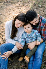 Fototapeta premium Picture of lovely parents hugging little baby toddler son, in autumn park or forest, sitting together and enjoying their family time, playing outdoors. Happy family in autumn nature