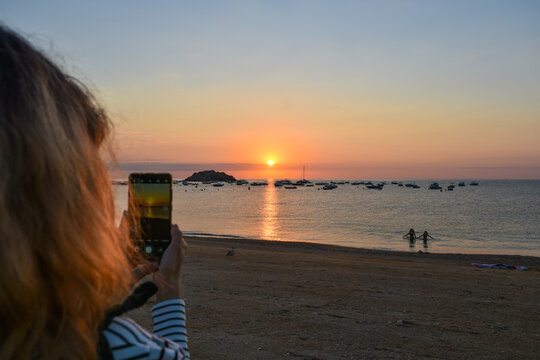 Hands of a woman taking photos with the mobile on the beach of Tossa de Mar during sunrise.