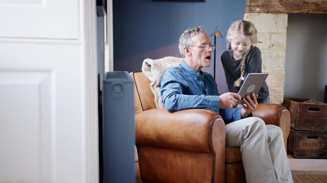 Grandfather and granddaughter at home sitting in armchair using digital tablet together 