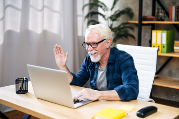 Handsome smiling older man with mustache and beard is using a laptop computer for online video call at home office. A gray-haired man is waving at webcam