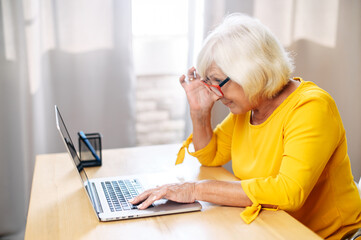 A senior concentrated business lady staring at the screen and lowered her eyeglasses while using...