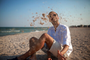 Young man sits on beach, has a fun and throws sand near sea.