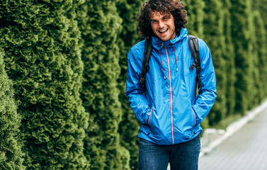 Horizontal outdoor shot of a young man student with curly hair with backpack smiling broadly walking to the college. Happy male walking in the street on a rainy day. Hipster guy has joyful emotion.