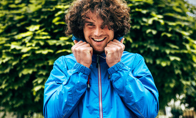 Portrait of a happy curly young man smiling broadly wearing blue raincoat during the rain outside. Positive male enjoying the rainy day hiding in the green leaves.