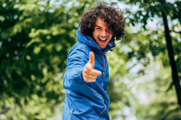 Positive young man smiling braodly in blue raincoat after the rain outdoor. Cheerful male enjoying the weather in the city street. The guy has joyful expression and showing thumbs up.