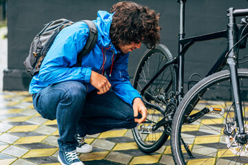 Obraz premium Side view of a man fixing with his bike after cycling in the street on a rainy day. A male courier with curly hair delivers parcel and fixing his bicycle.