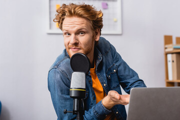 curious redhead announcer looking at camera while pointing with hand near microphone and laptop