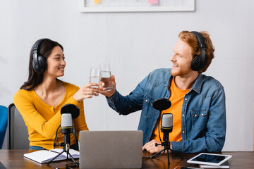 interracial couple of young radio hosts clinking glasses of water at workplace
