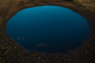 large geometrically smooth round blue water puddle with sky reflection in pebbles of gravel road