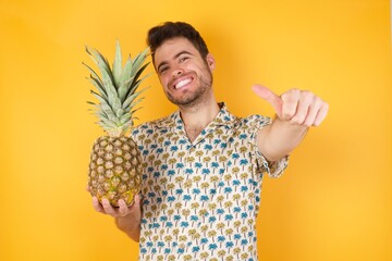 Young man holding pineapple wearing hawaiian shirt over yellow isolated background  approving doing...