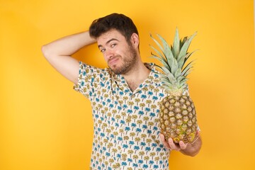 Young man holding pineapple wearing hawaiian shirt over yellow isolated background  being confused...