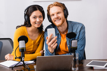 young broadcaster holding smartphone while looking at camera together with asian colleague in radio...
