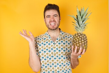 Indignant Young man holding pineapple wearing hawaiian shirt over yellow isolated background gestures in bewilderment, frowns face with dissatisfaction, supper. Negative emotions and facial expression