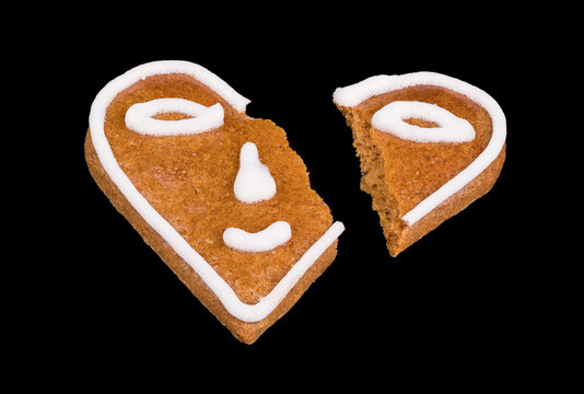 Broken heart from baked gingerbread decorated by smiley isolated on a black background. Close-up of damaged sweet cookie. Smiling emoticon divided into two parts. Symbolic end of love or relationship.